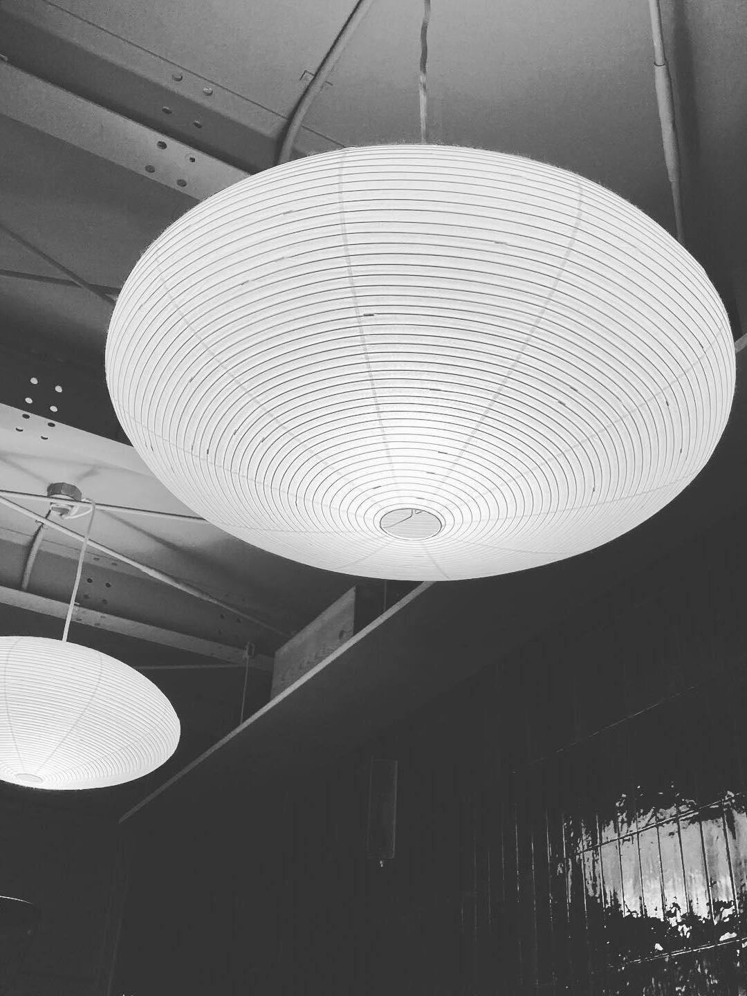Black-and-white view from below a broad ribbed paper lantern under exposed beams, with another lantern behind
