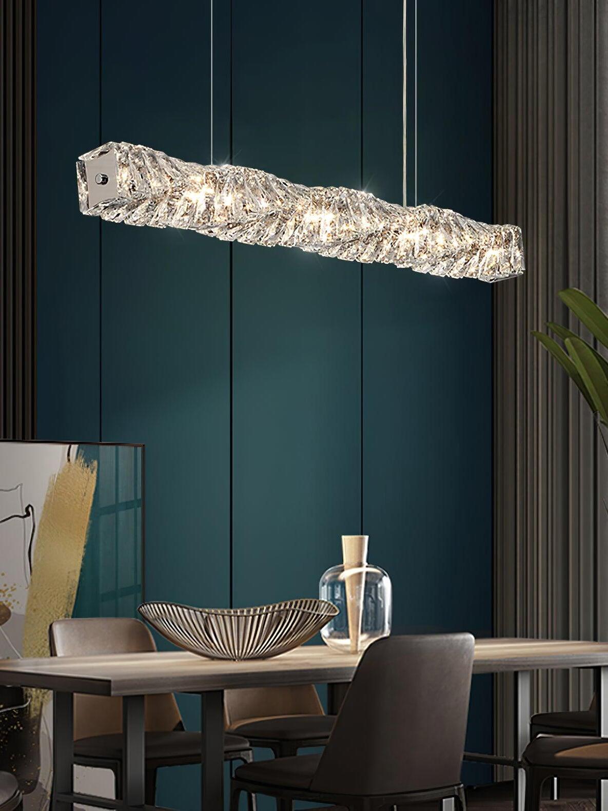 Linear crystal pendant light above a dining table with a bowl and glass jug against a teal panelled wall