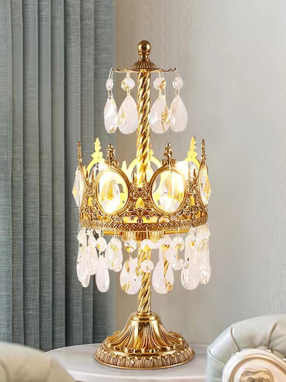 Illuminated crystal table lamp with upper drops beside grey curtains and the edge of a pale chair.