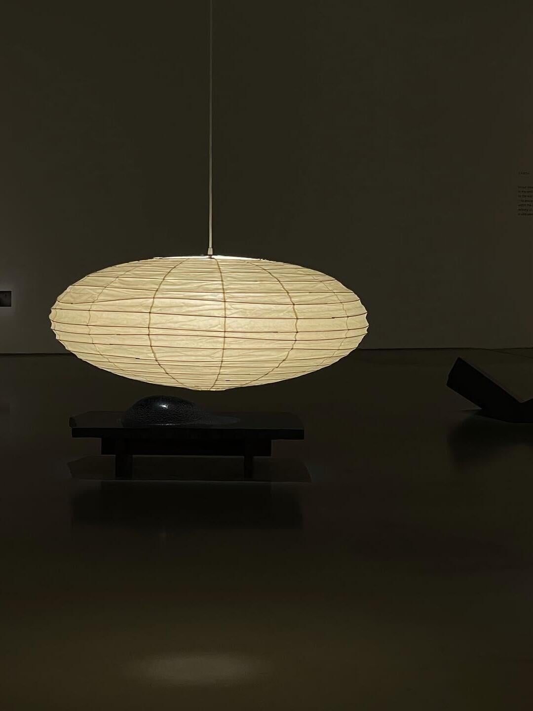 Illuminated flattened paper lantern above dark sculptural furniture, with horizontal ribs and vertical supports visible