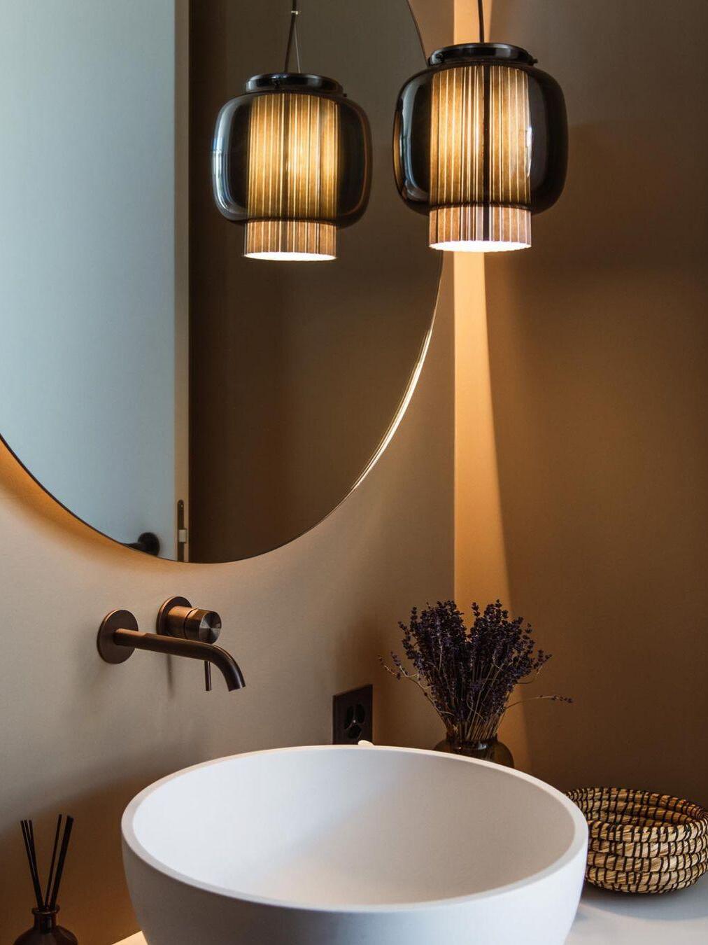 Lit smoke-grey Manila T pendant and its mirror reflection above a white sink beside dried flowers