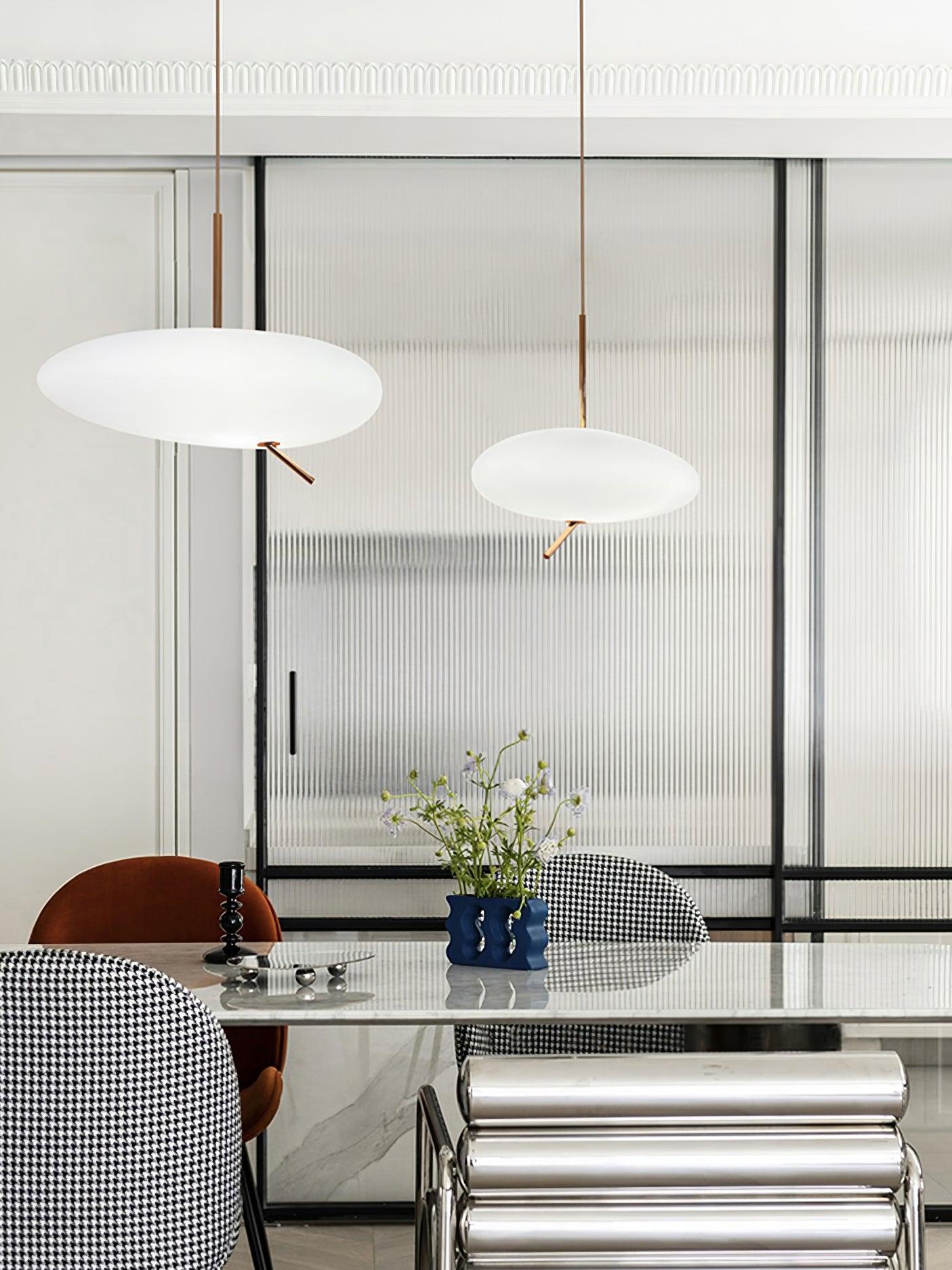 Two white pendant shades above a dining table with ribbed metal chairs and a blue vase