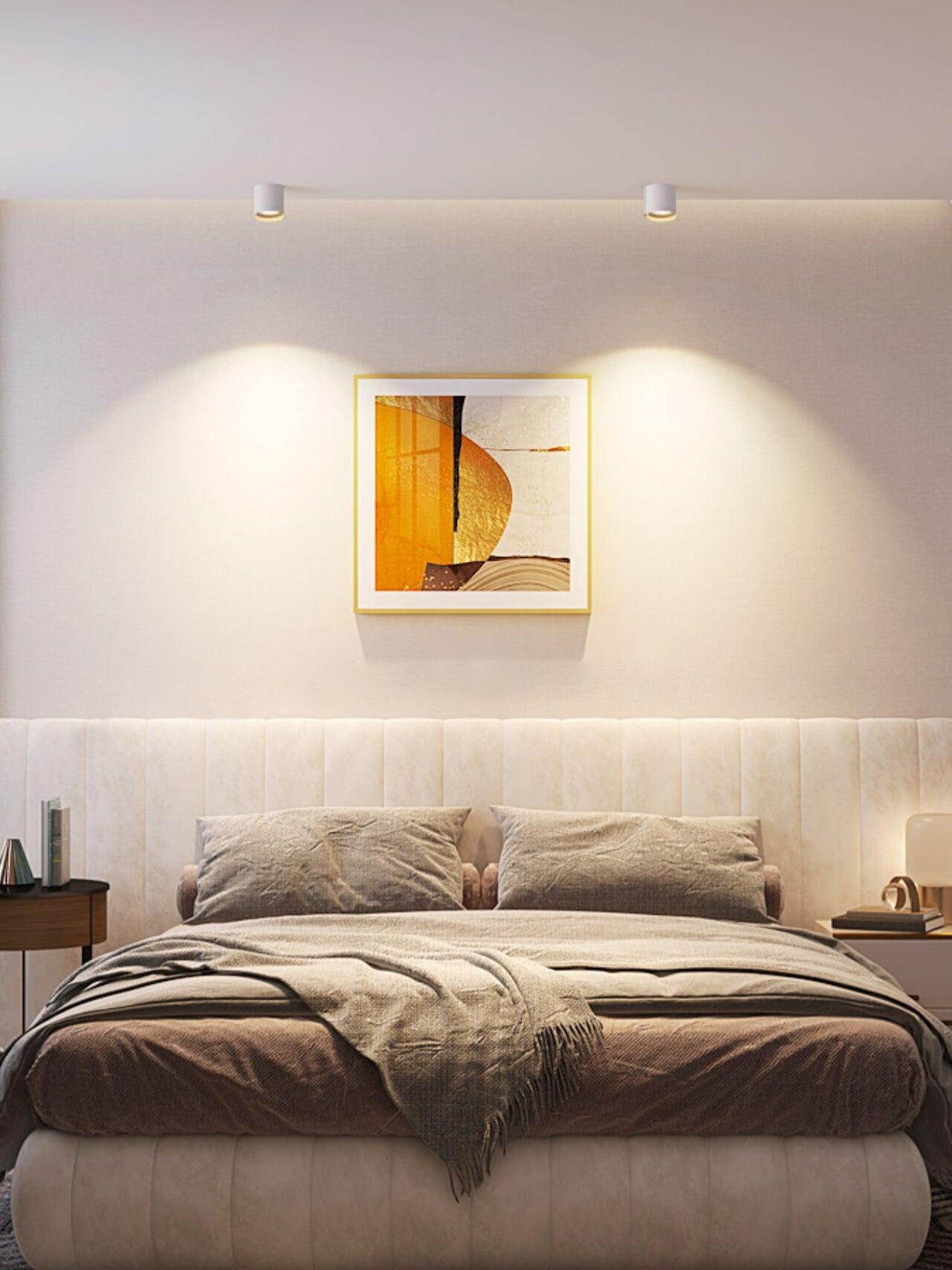 Two white ceiling lights above a pale upholstered bedhead and gold-framed abstract artwork