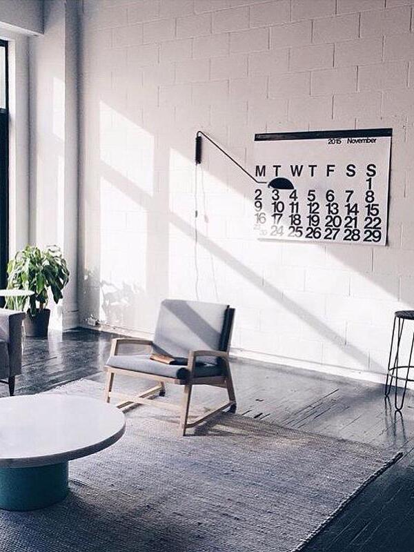 Black dome wall light above a grey reading chair beside a large calendar on white brick, with a round low table