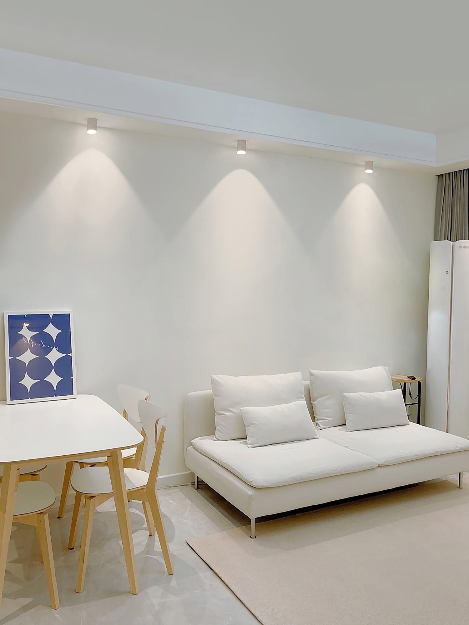 Three white ceiling lights along a beam above a white sofa and dining area, with blue framed print and curtains
