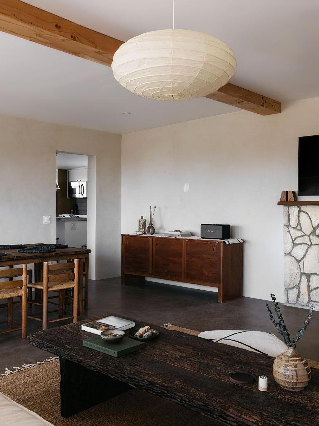 Broad white paper lantern beneath a timber beam in a living and dining room with wooden furniture and stone fireplace