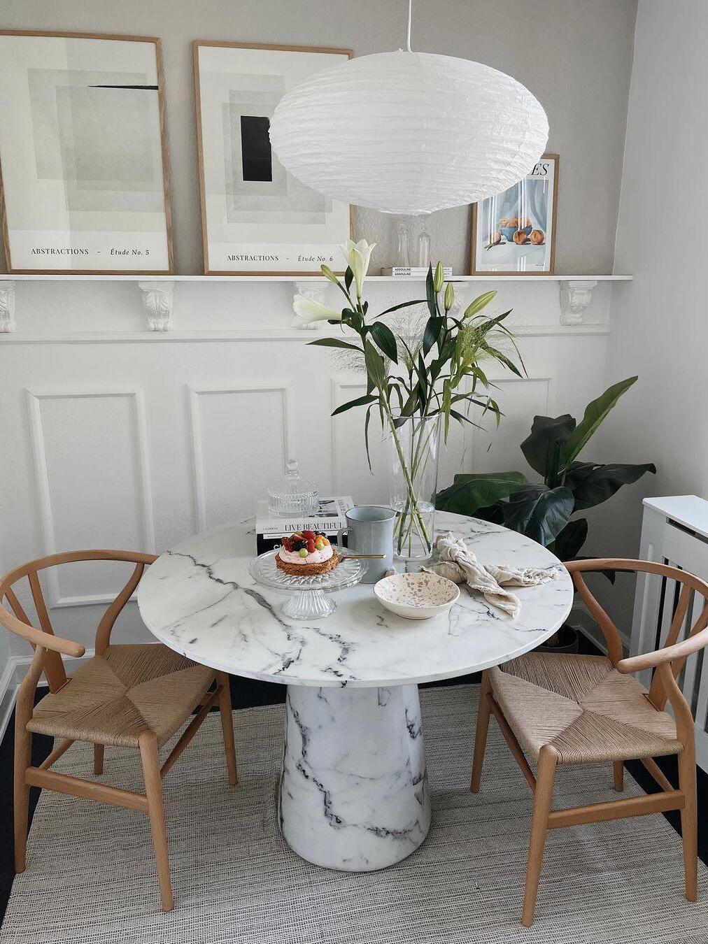 White textured paper lantern over a round marble-look table with lilies, dessert and woven chairs, with framed prints behind
