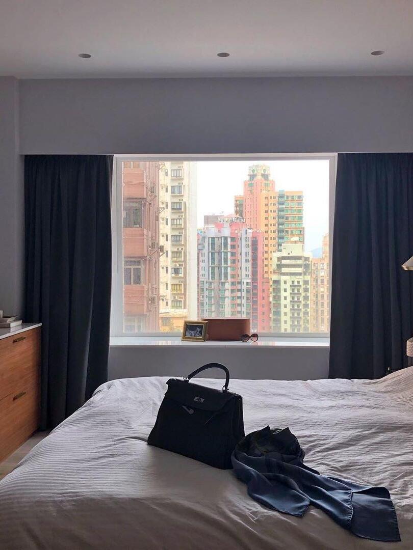 Bedroom scene with a city-view window, dark curtains, a handbag and clothing on a white bed