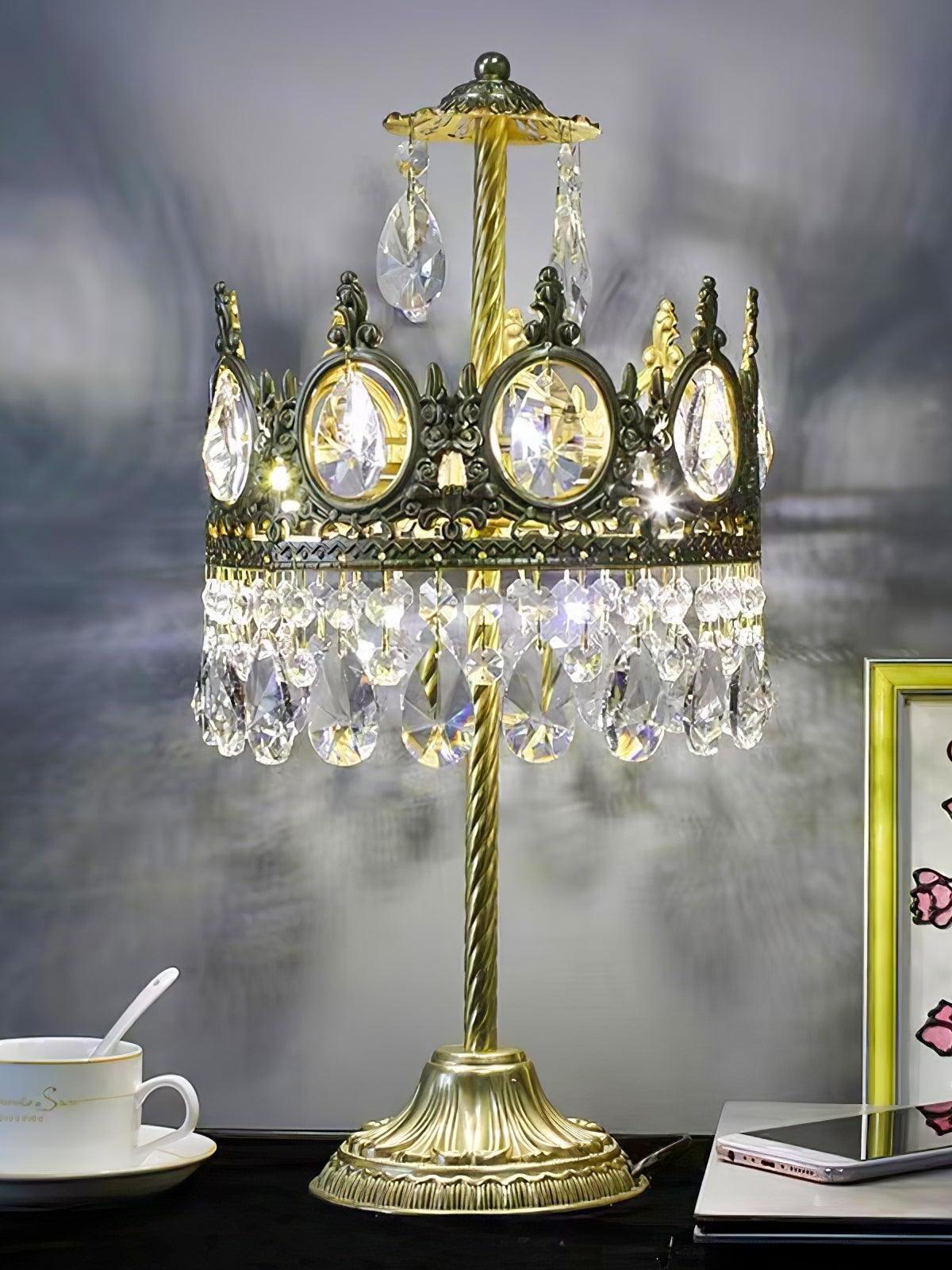 Lit crystal table lamp with upper drops beside a cup and books, casting patterns on a grey wall.