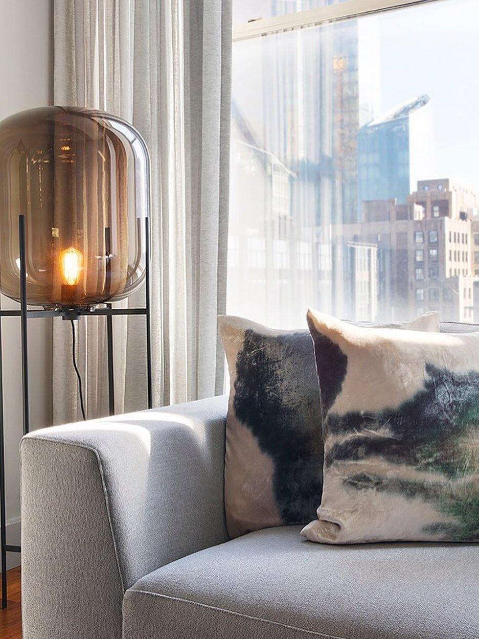 Grey glass floor lamp with a glowing inner bulb beside a window sofa.