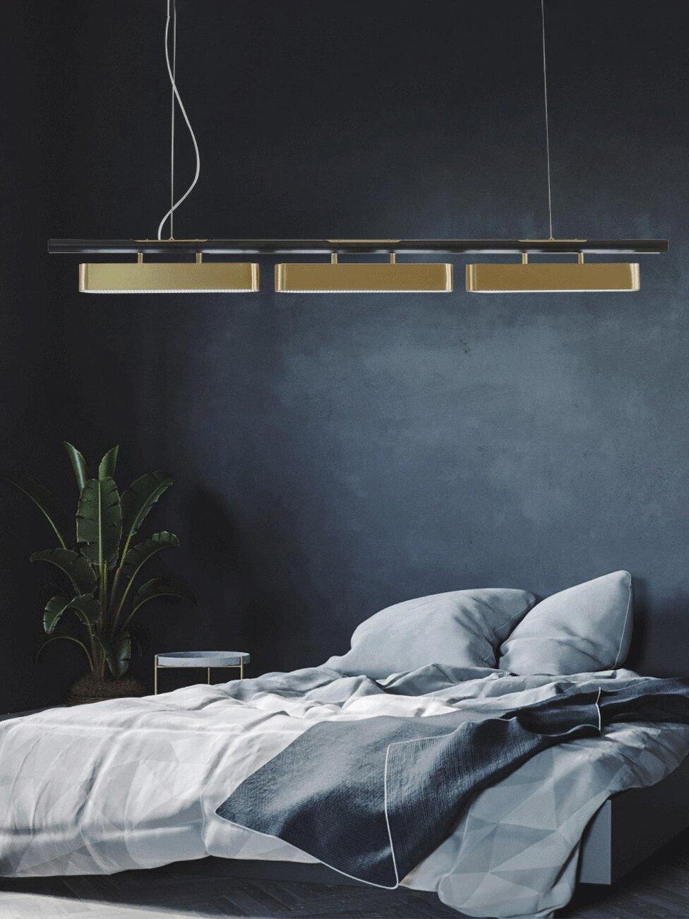 Colt three-module brass-and-black pendant above a bed with blue-gray pillows and a blanket beside a plant