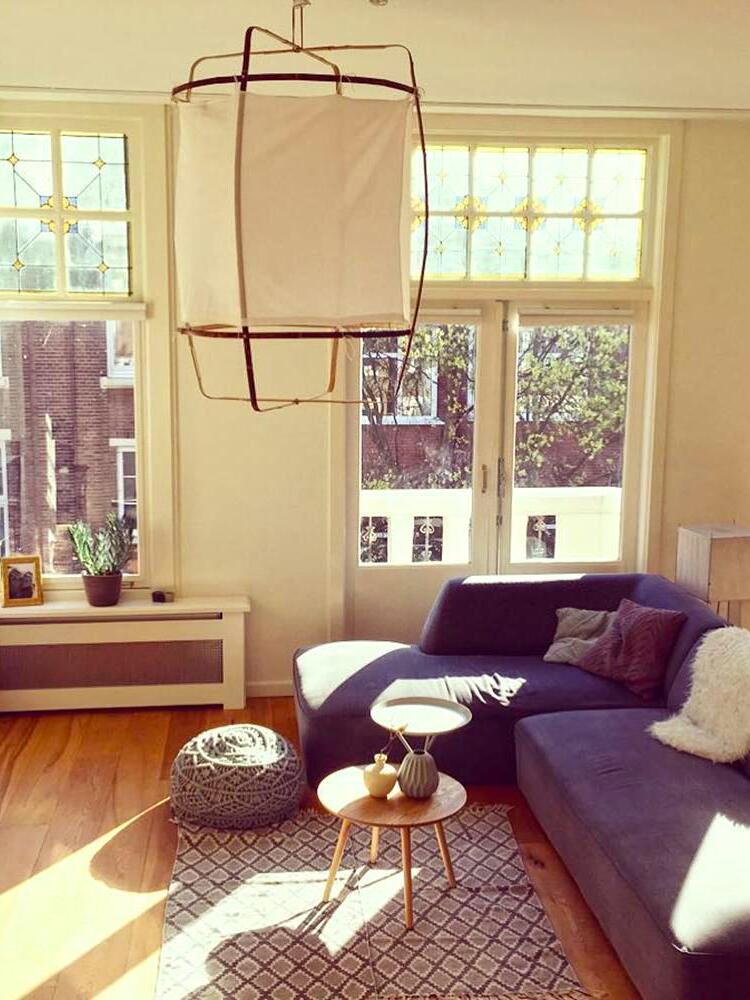 White bamboo lantern pendant above purple sectional sofa, wooden floor and small round tables near tall windows