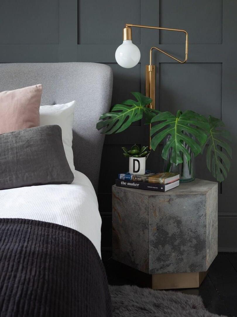 Brass-tone bent-arm wall lamp with white globe bulb beside a bed and table with books and green leaves
