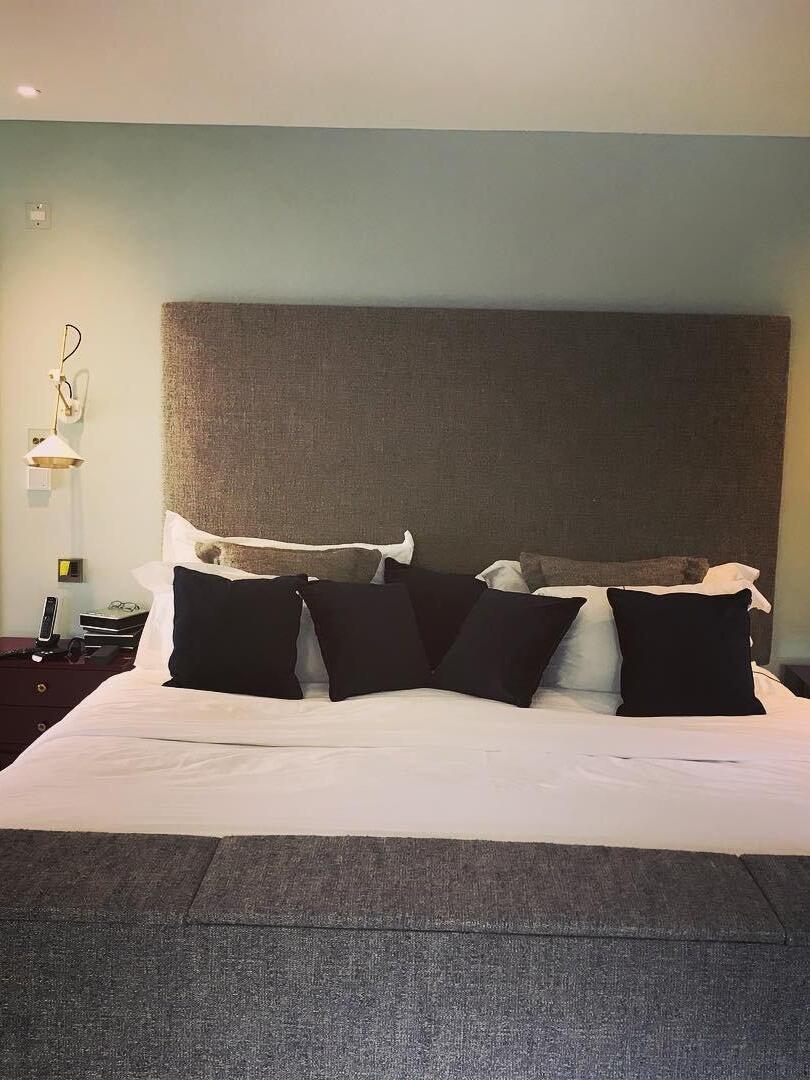 Bedroom with tall brown headboard, dark cushions and a white cone wall light at the left edge