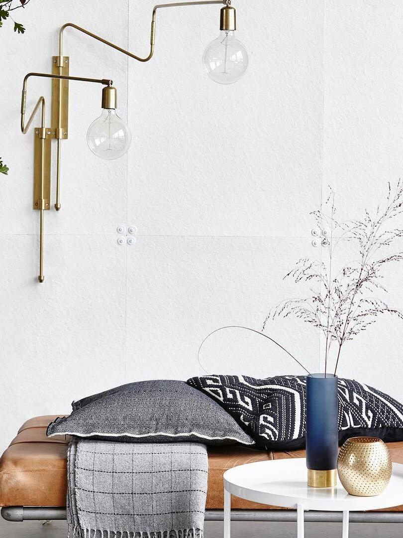 Two brass-tone bent-arm wall lamps above a brown sofa and white table with a tall blue vase