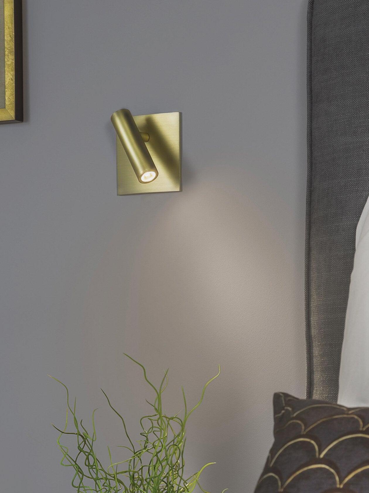 Gold-tone Enna lit cylindrical wall light on grey wall above plant, beside upholstered headboard and framed picture edge