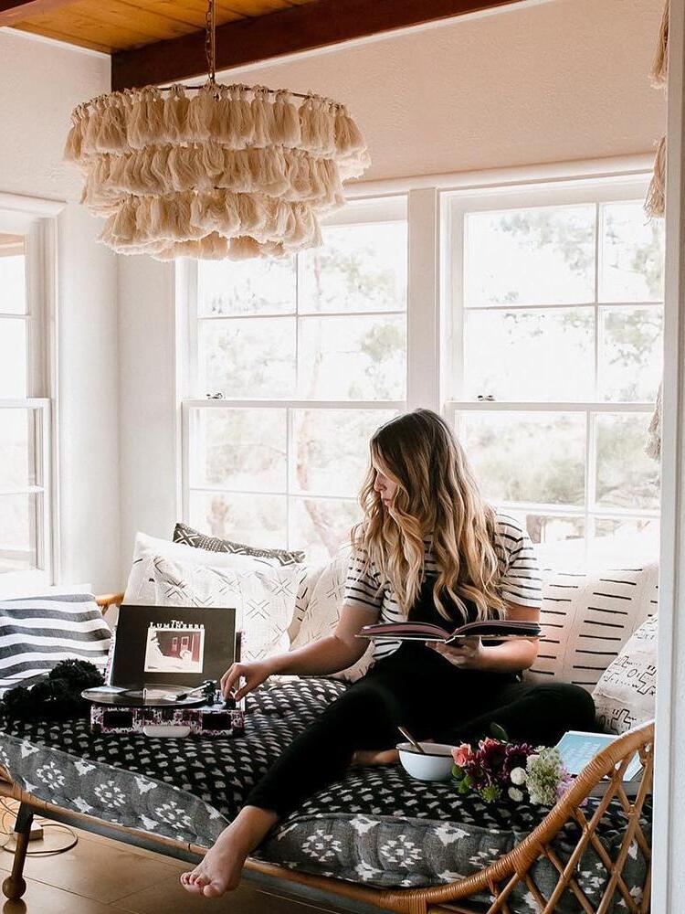 Cream layered tassel chandelier above a cane daybed and a reader beside bright windows and wood ceiling beams