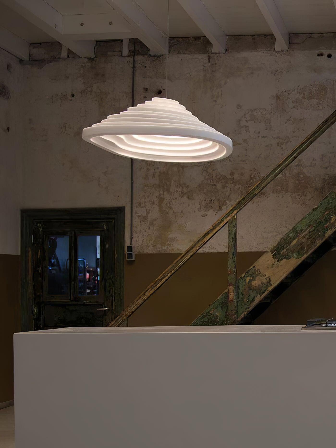 Lit stepped pendant over a pale counter with a staircase and textured wall behind.