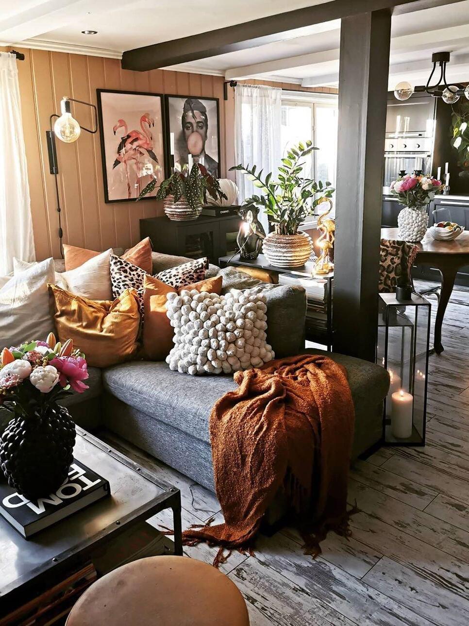 Black bent-arm wall lamp above a grey sofa in a plant-filled room with orange cushions and artwork