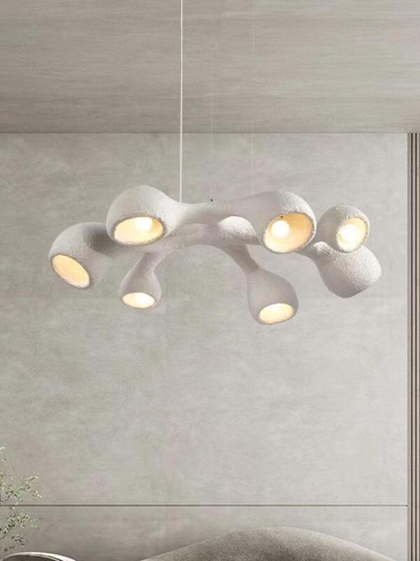 White looped branching pendant before a pale wall panel above a cropped sofa and small plant