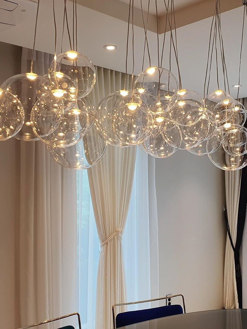Clear glass globes with lit inserts above a dining area beside a curtained window and blue chair