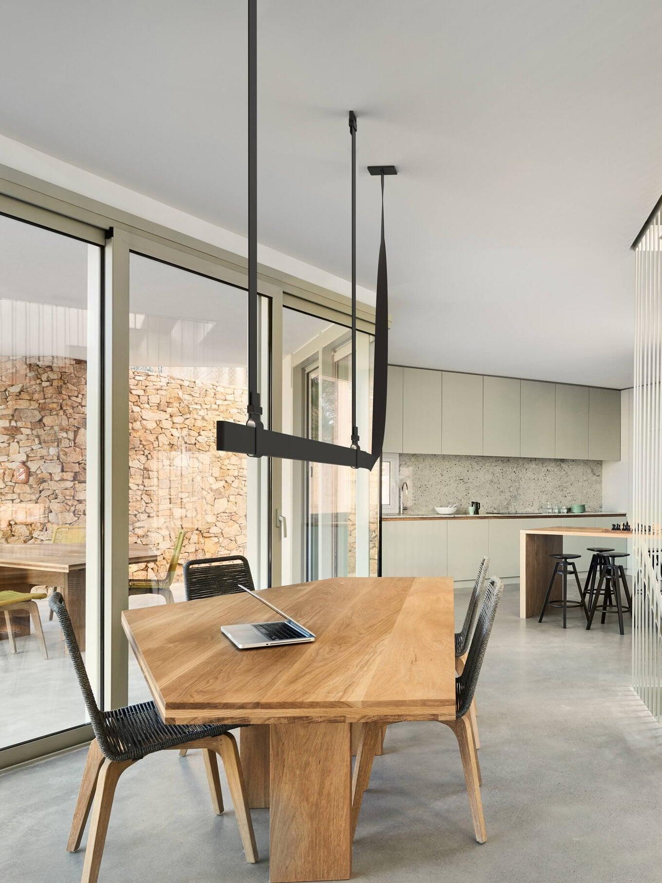 Oblique view of a black belt-shaped pendant above a wood table with a laptop beside a large window