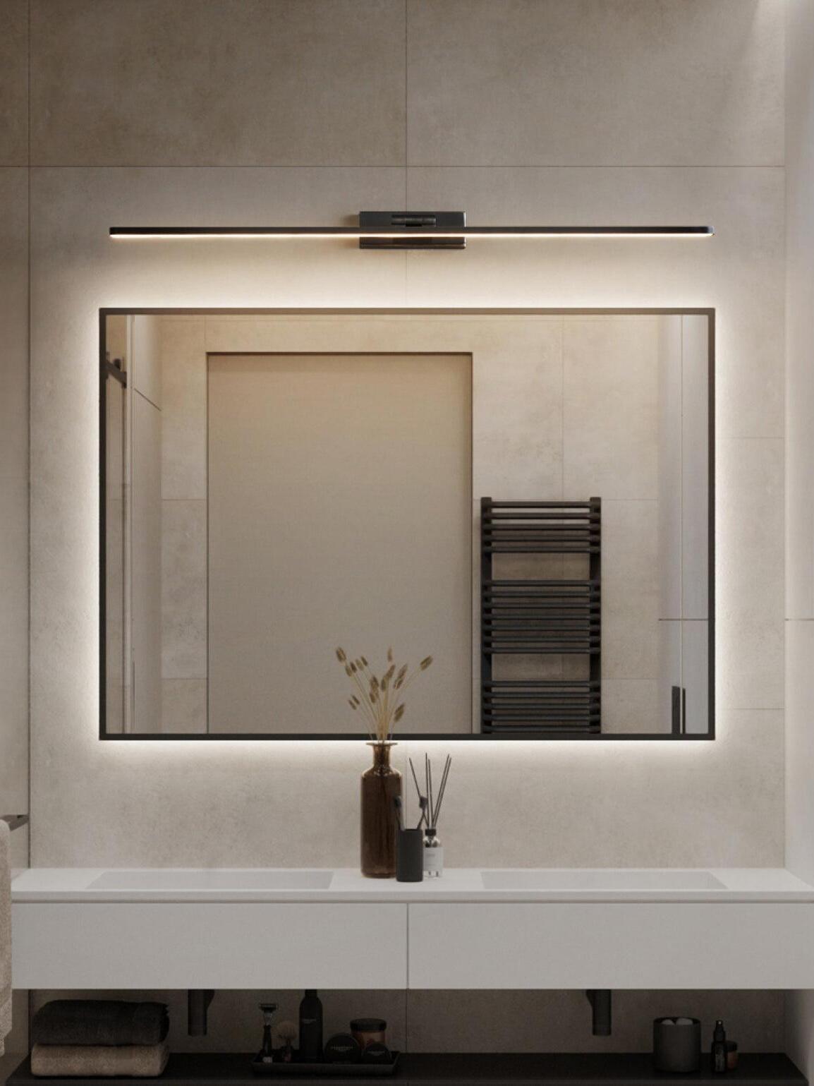 Black linear wall light above black-framed backlit mirror and white double-basin cabinet against tiled wall
