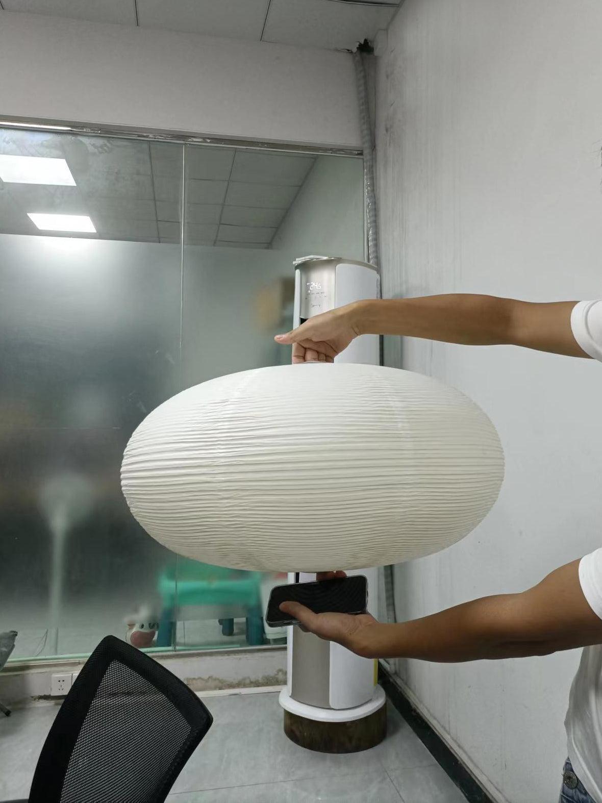 Person holding a broad flattened white ribbed paper shade in a workspace, with a phone held below
