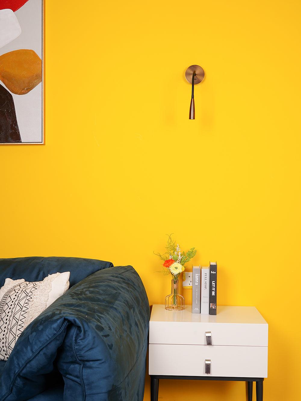Bronze-toned Flexi facing forward with its head down on a yellow wall above a cabinet, flowers and books