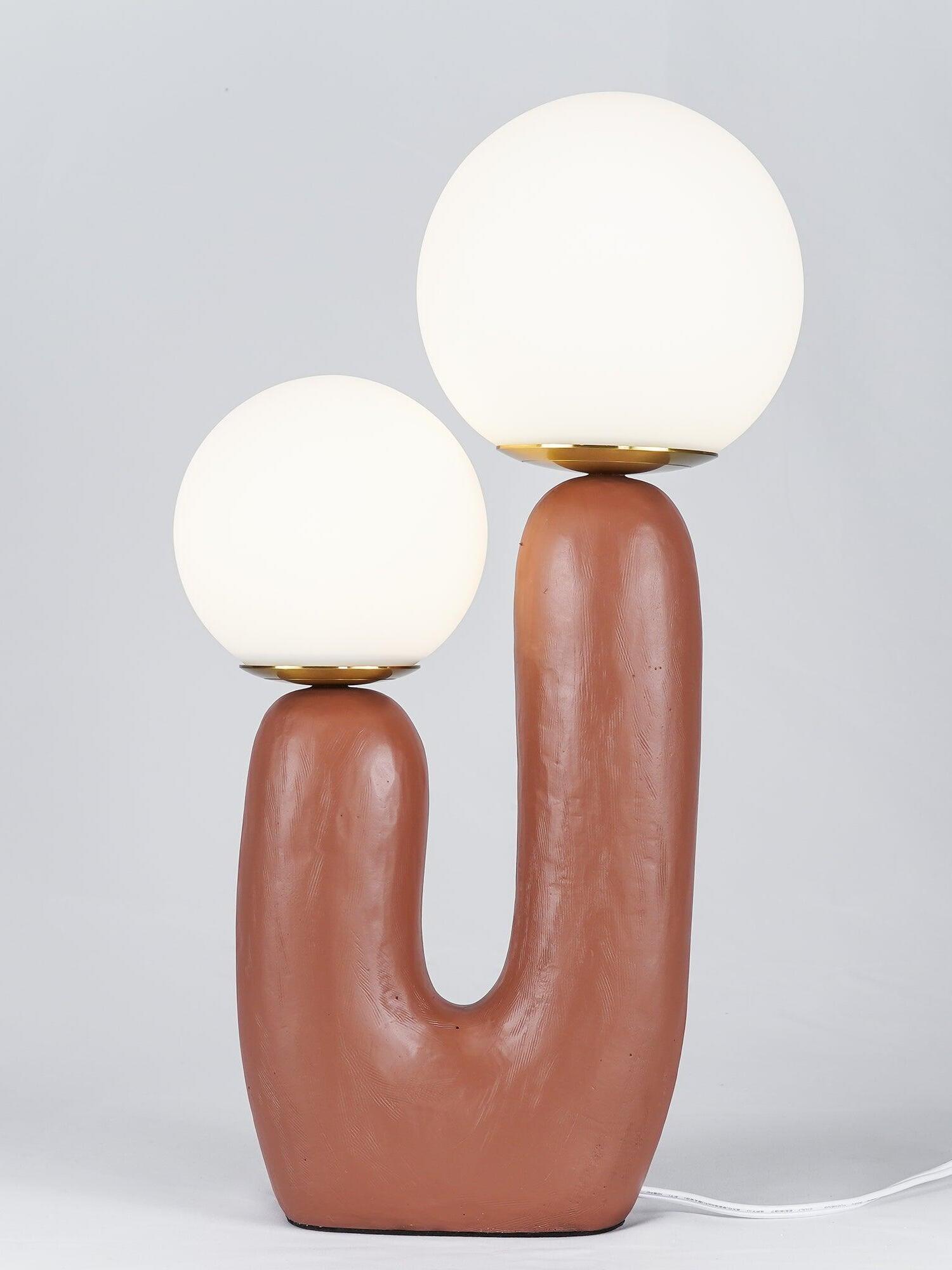 Close view of terracotta Oo Rough lamp against grey, two glowing opal globes above its textured U-shaped body