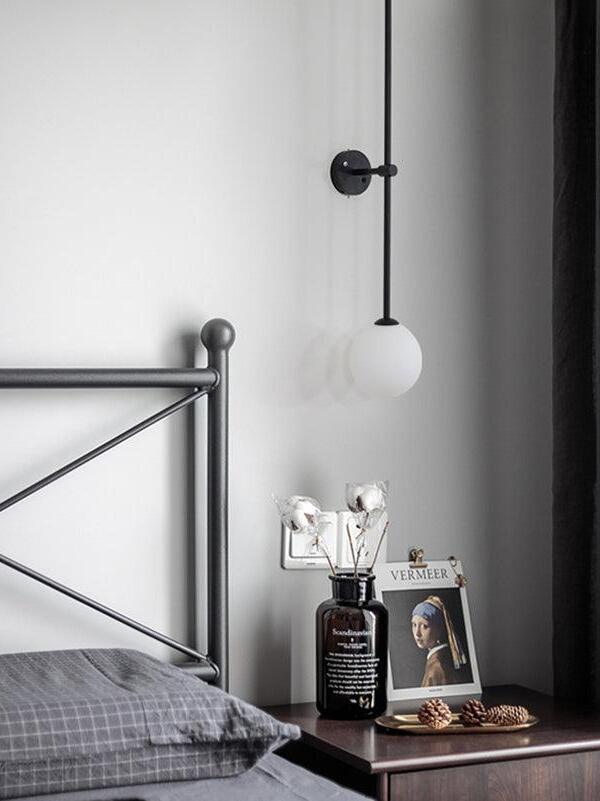 Unlit white globe and black rod beside a metal bedframe, with a vase, book and pine cones on the table