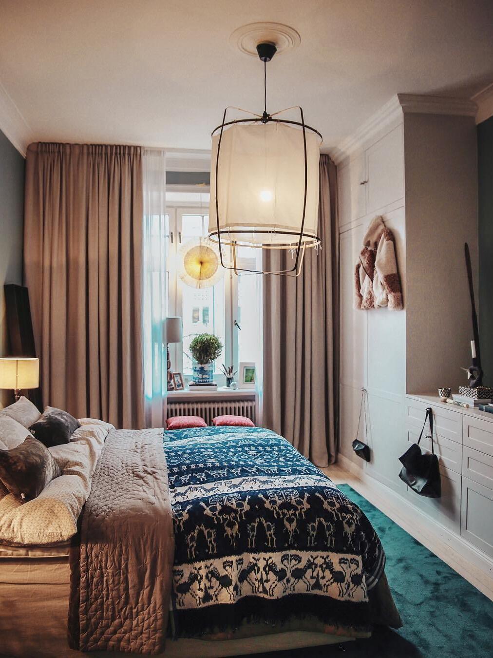 White lantern pendant glowing above a bed with patterned blue cover, beige curtains and radiator below the window