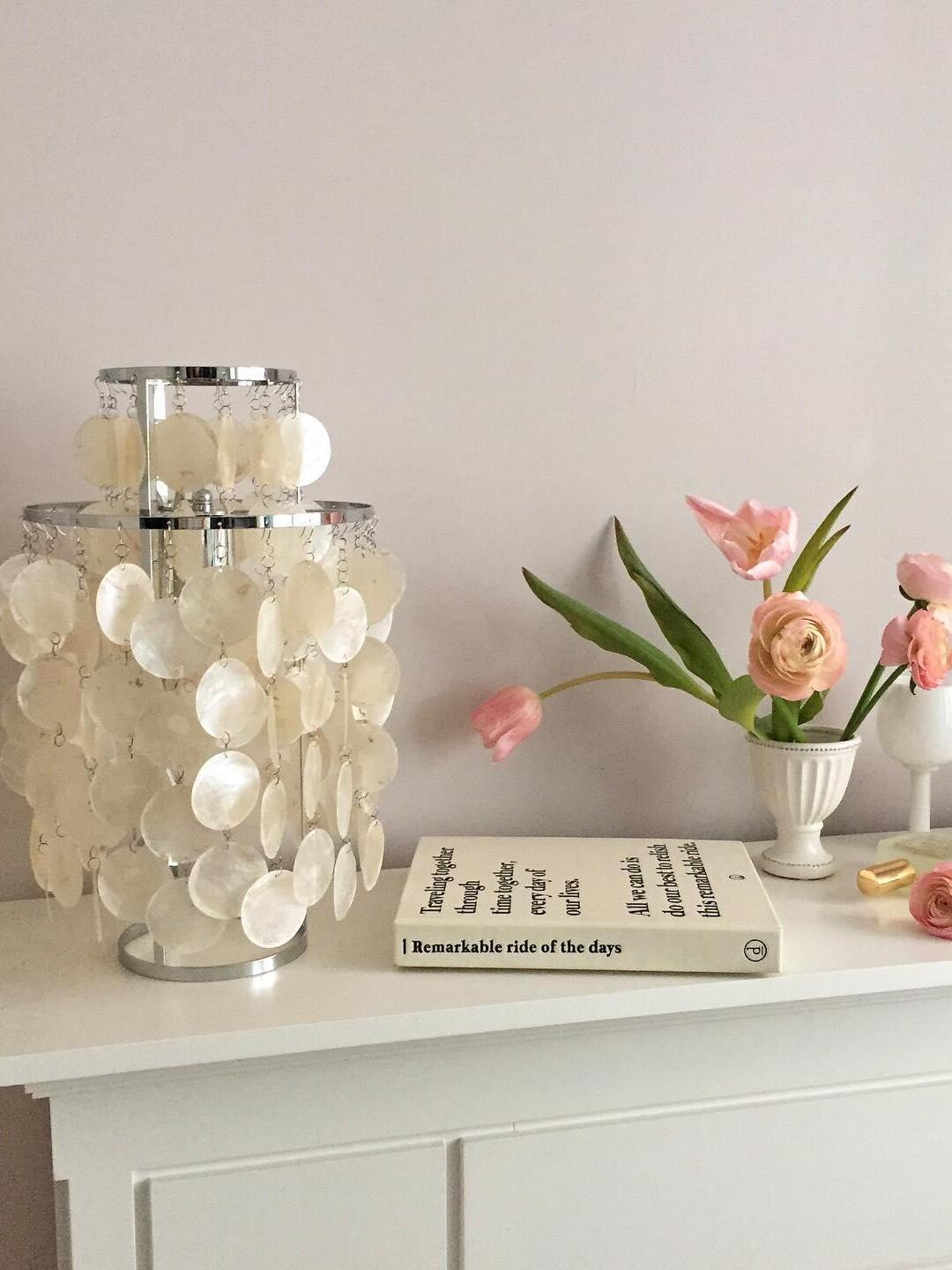 Unlit shell table lamp on white cabinet beside pink flowers