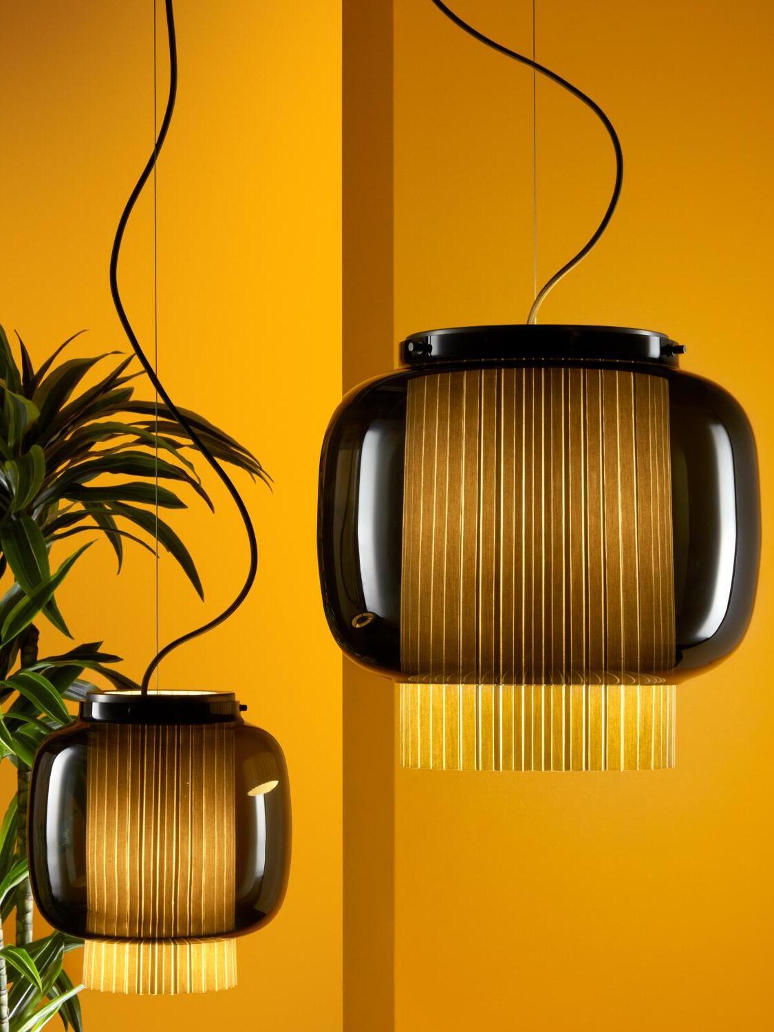 Two different-size Manila T pendant lights with smoke-grey glass against a yellow wall beside a green plant