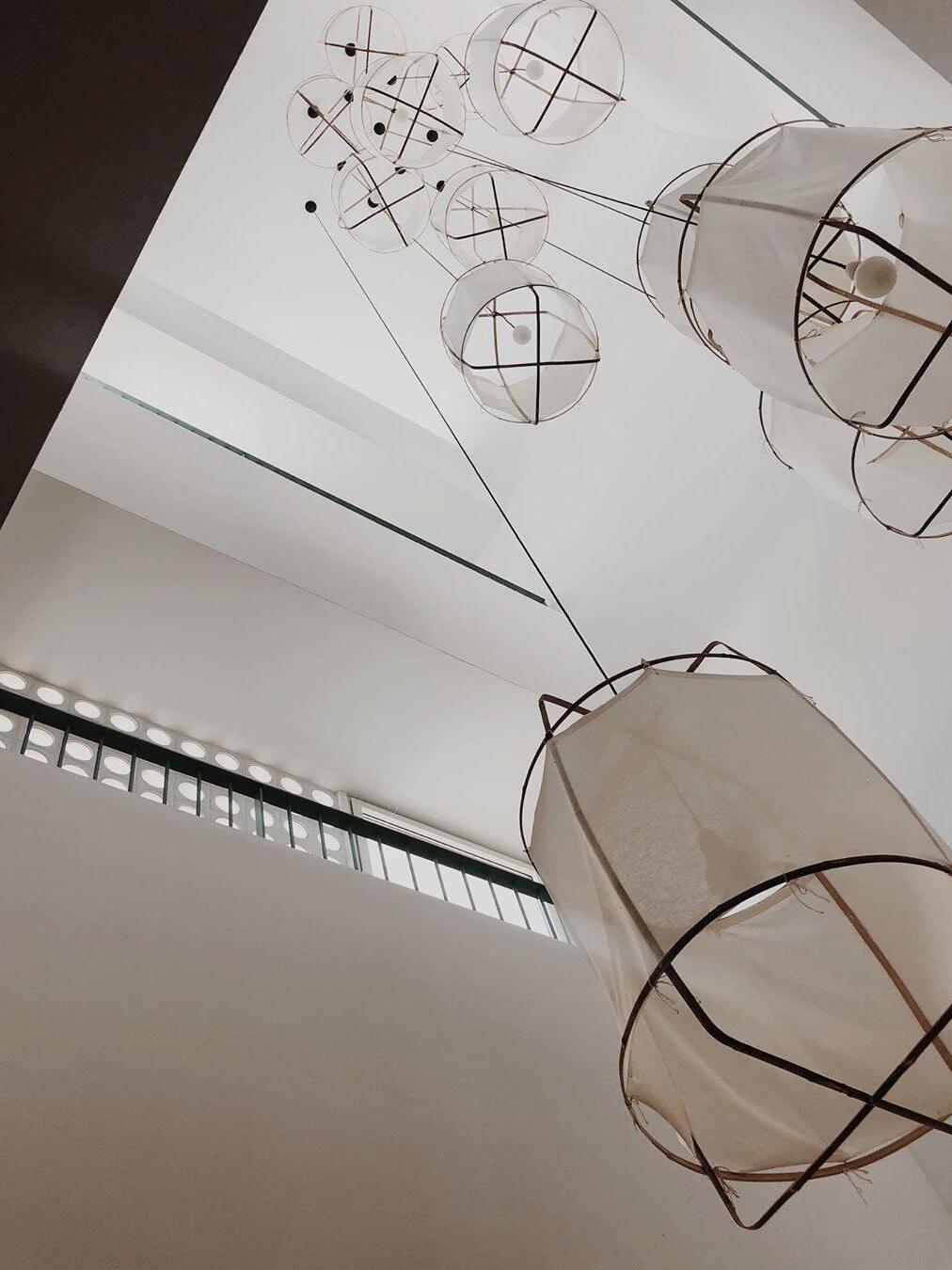 Upward view of many bamboo pendant frames and fabric sleeves with visible bulbs in a tall white gallery