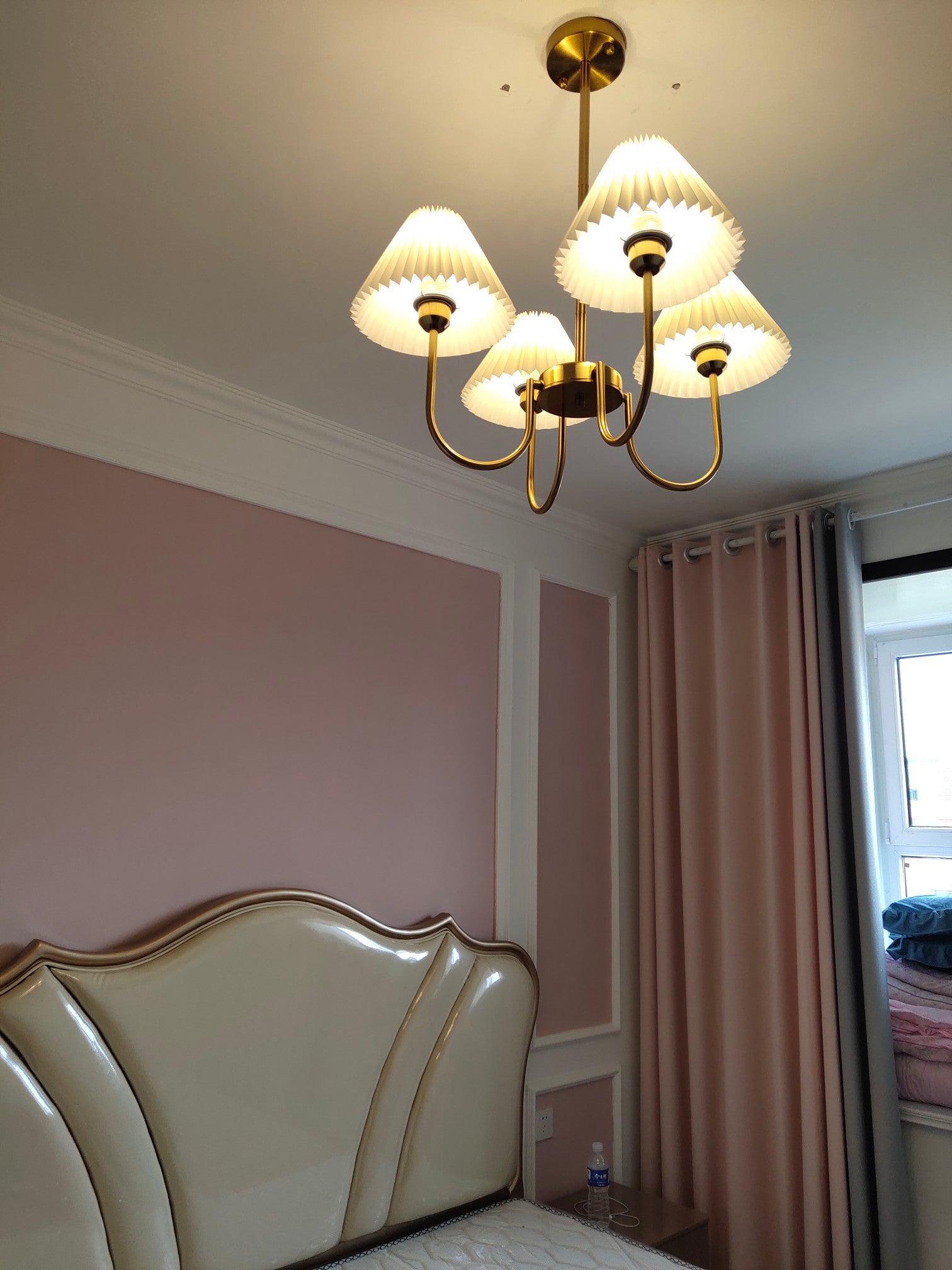 Low-angle view of a lit pleated-shade chandelier above a curved headboard beside pink curtains