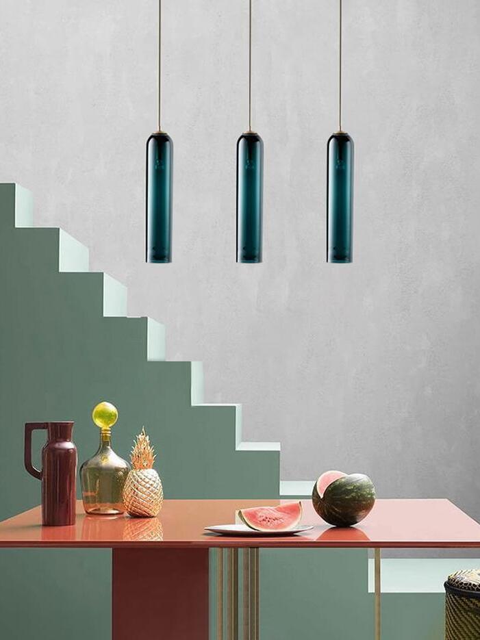 Three blue-green glass tube pendants above a coral dining table with colourful vessels and fruit