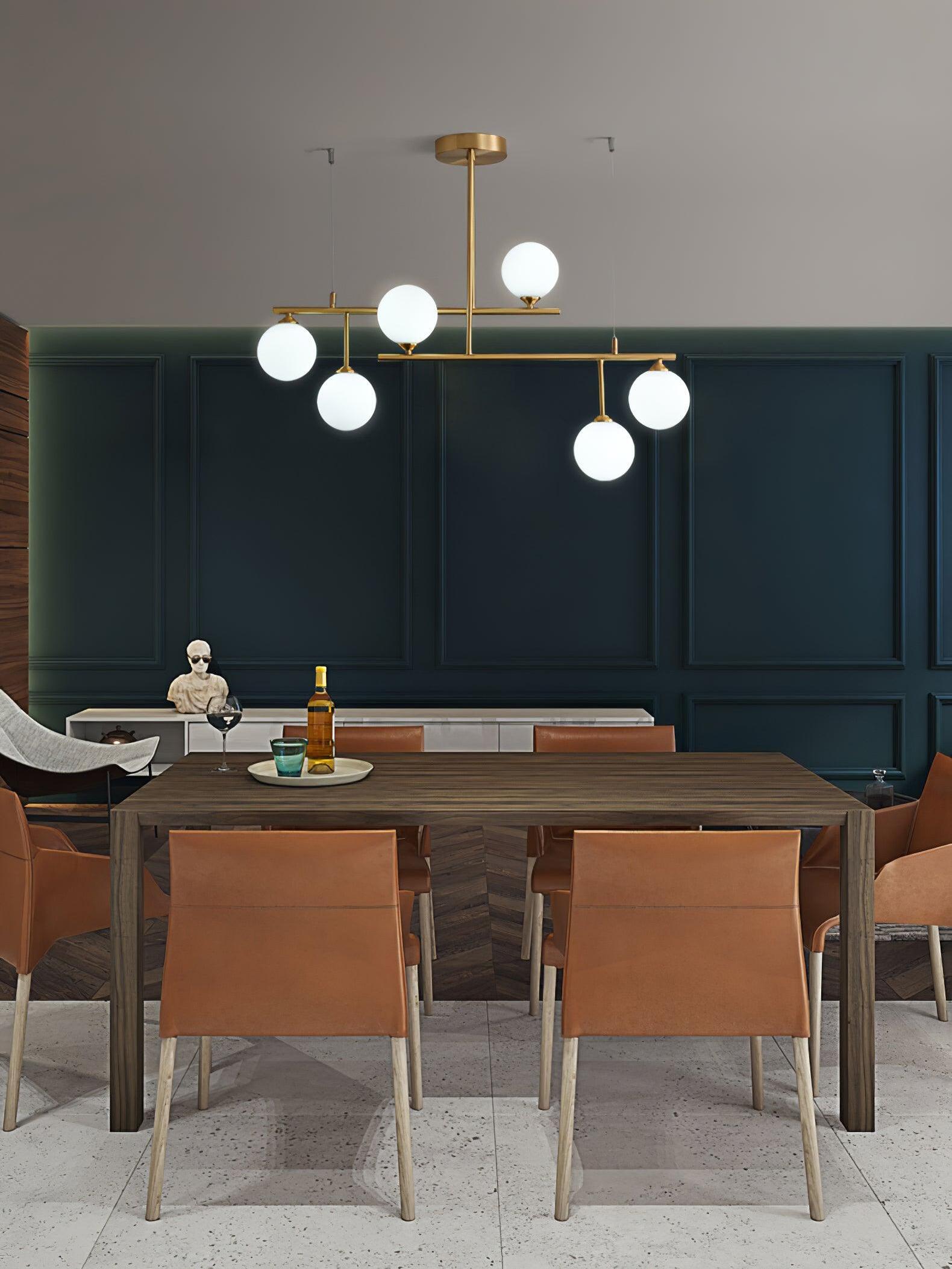 Brass-tone Arteriors chandelier above a wooden dining table and tan chairs in a green-panelled room