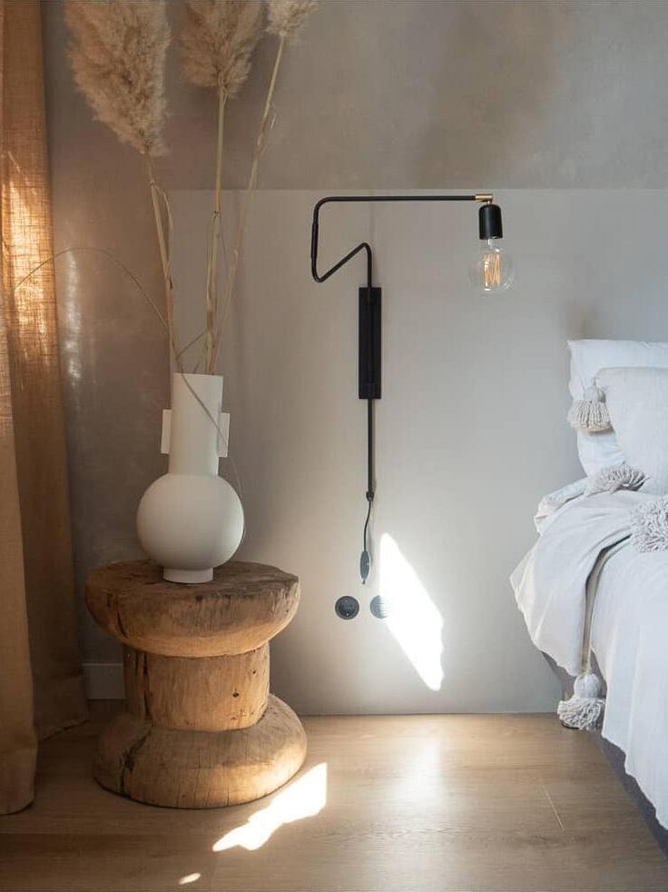 Black bent-arm wall lamp with inline switch beside white bedding and a wooden spool table