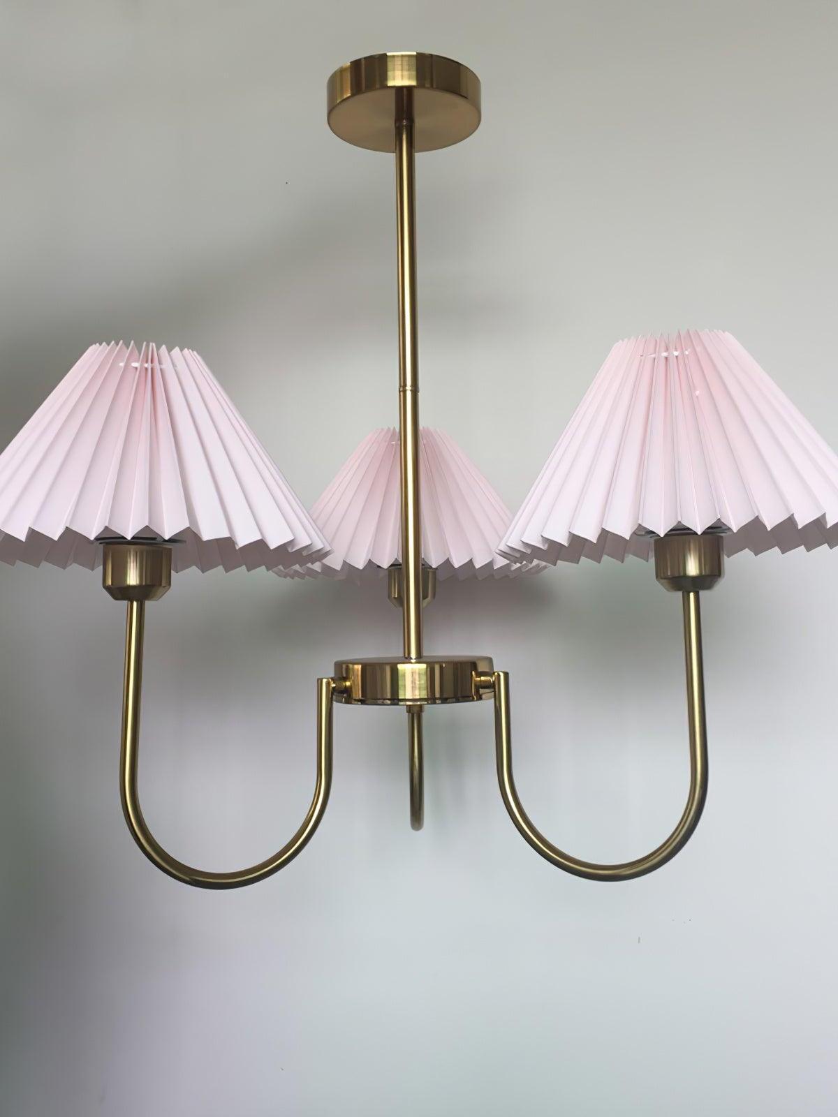 Three pleated shades appearing pink on curved gold-tone chandelier arms against a grey wall