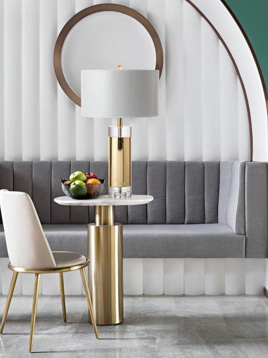 Gold-tone column lamp on round marble-look table before grey banquette and mirrors