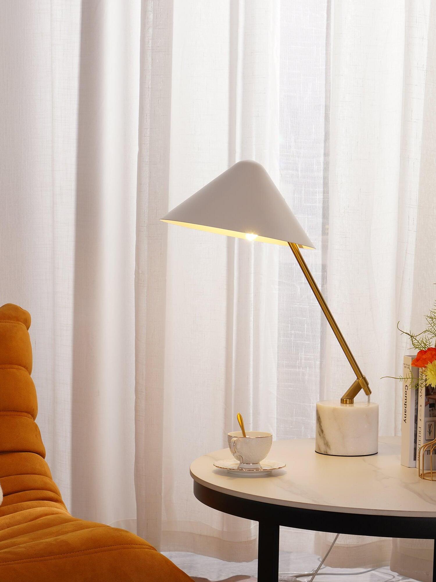 White angled-shade lamp on a round table beside a cup, books and orange chair