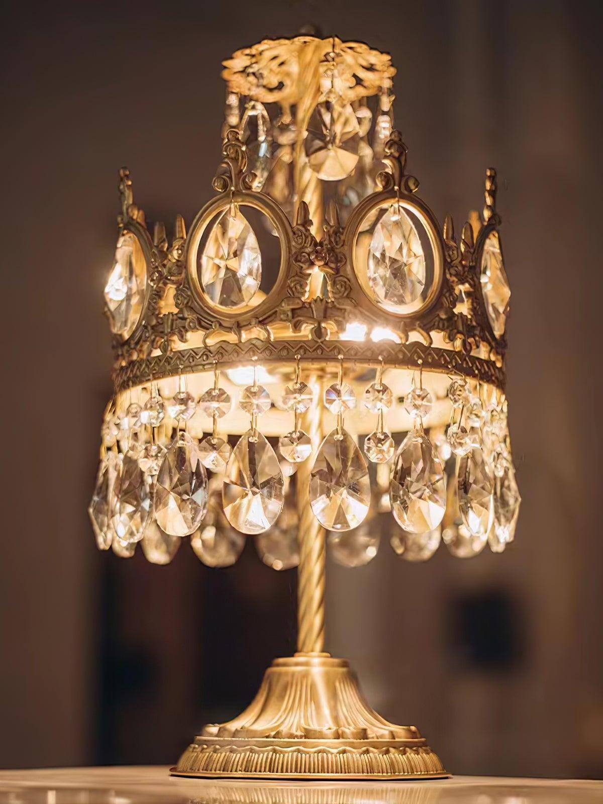 Warmly lit crystal table lamp with hanging drops above and below its ornate circular crown.