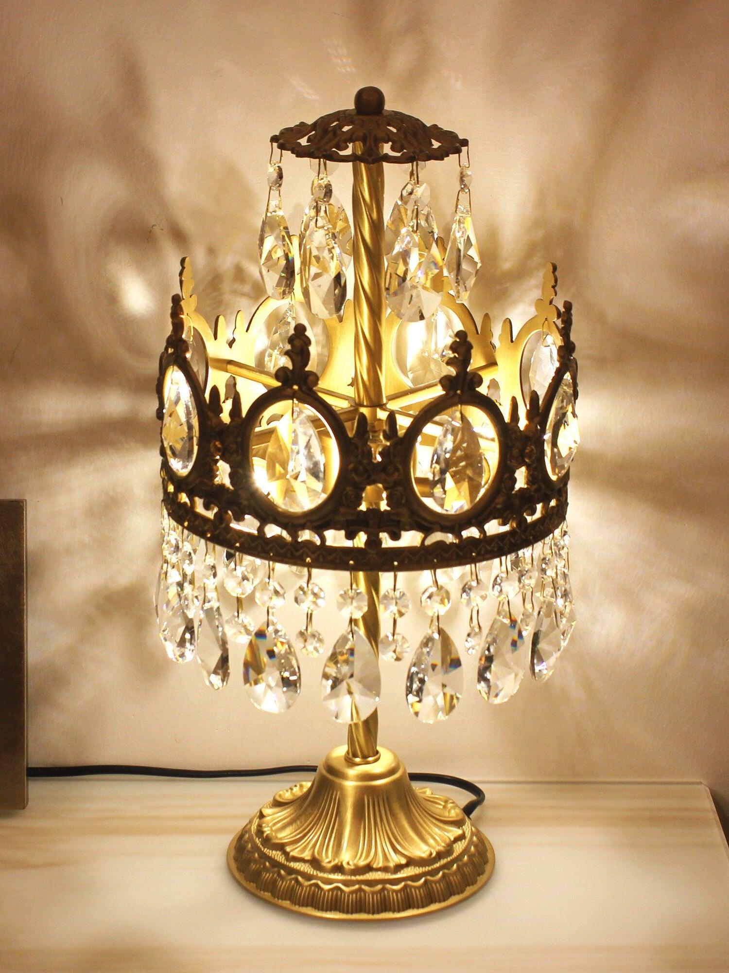 Lit crystal table lamp with an upper drop tier and decorative crown, casting patterns on a pale wall.