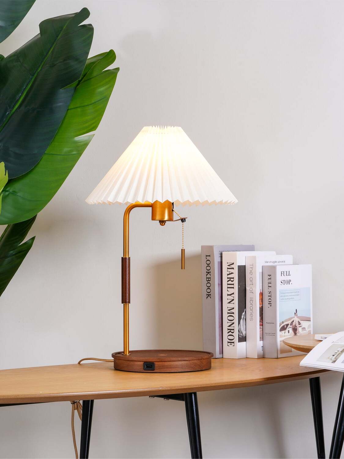 Lit pleated table lamp with a curved gold-tone upper stem beside upright books and green leaves