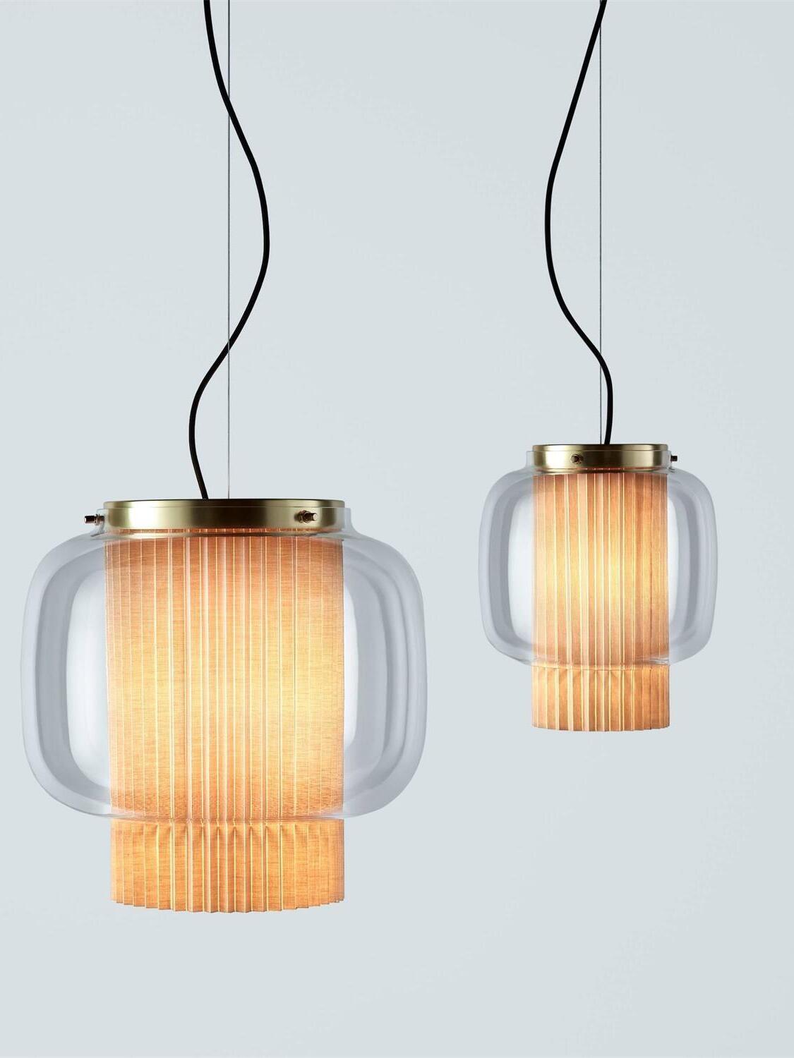 Two different-size Gold and clear-glass Manila T pendants with glowing pleated fabric diffusers against pale blue-grey