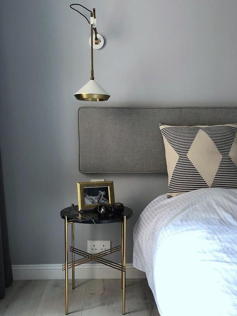 White cone wall light on a vertical arm above a gray headboard and a round black-and-gold bedside table