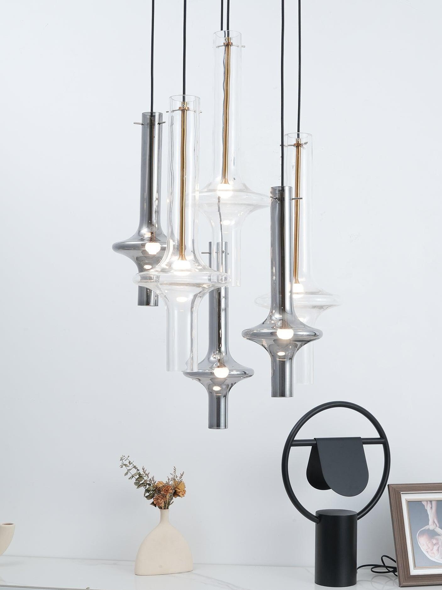 Six clear and smoky tube-shaped glass pendants over a white console with dried flowers, black table lamp and framed picture