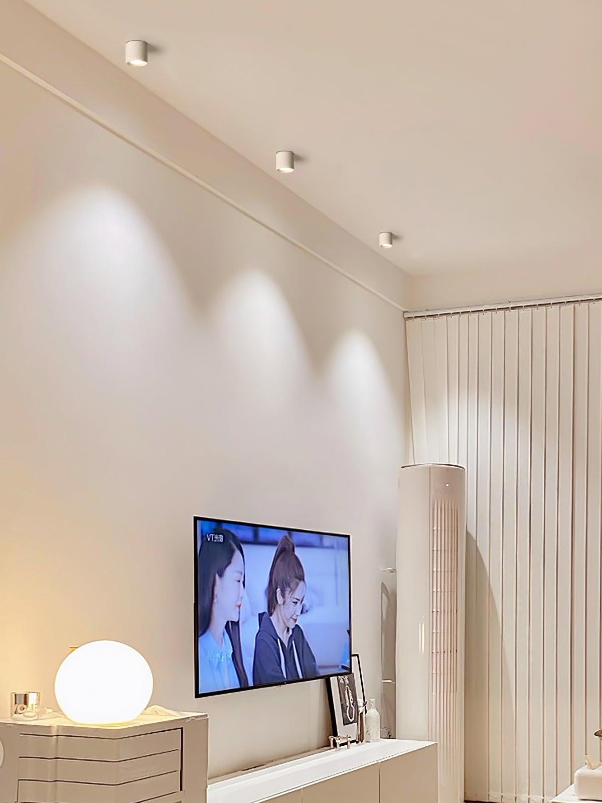 Three white ceiling lights along a TV wall, with round table lamp, white cabinet, tall air conditioner and vertical blinds