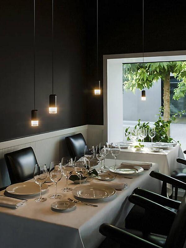 Three lit black pendant heads over a set dining table beside a bright window